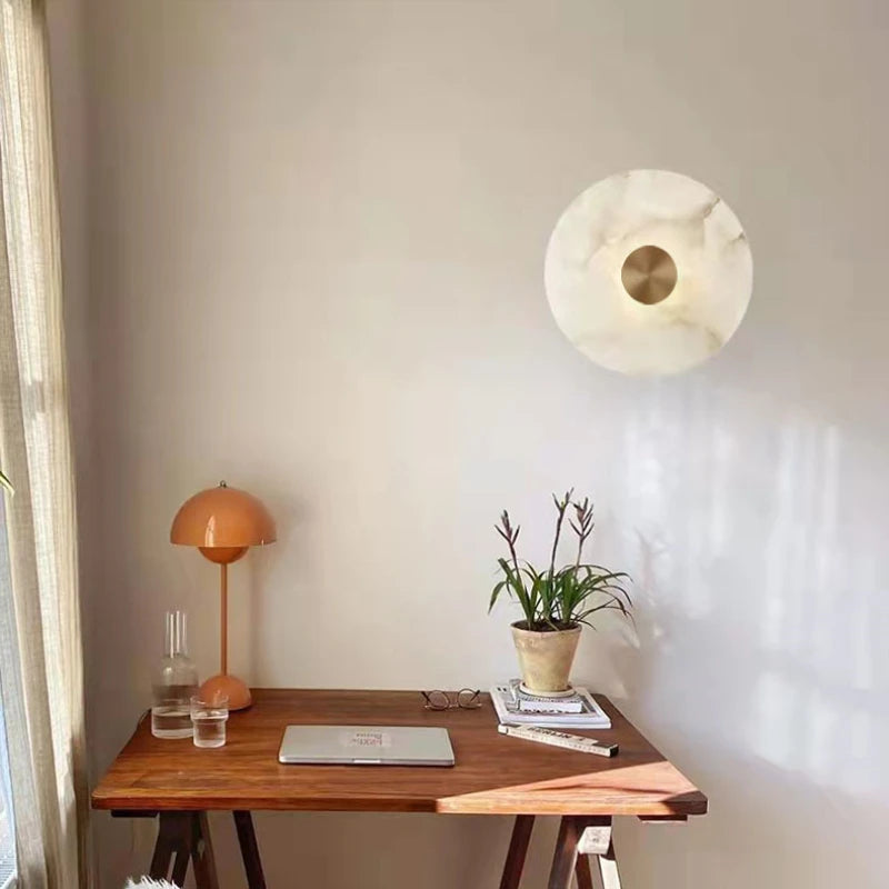 Wooden desk with a laptop, lamp, and plant against a white wall with a decorative light fixture.